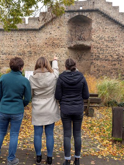 Drei Personen stehen vor einer alten Stadtmauer und schauen nach oben. Eine Person zeigt auf etwas an der Mauer.