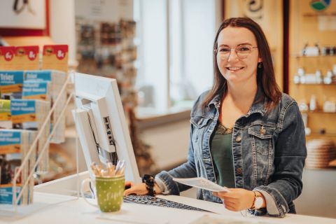 Eine Frau steht an einem hohen Schreibtisch mit Computer und Tastatur vor sich und lächelt in die Kamera. Hinter ihr ist der Souveniershop und einige Prospektregale der Tourist-Information.