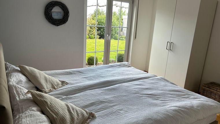 Une chambre lumineuse avec un grand lit et deux oreillers. Il y a de grandes fenêtres avec vue sur un jardin verdoyant.