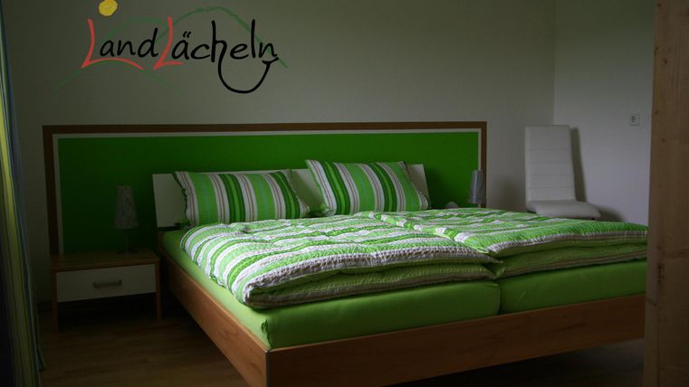 Ein modernes Schlafzimmer mit einem großen, grünen Bett und gestreiften Kissen. Der Raum ist hell und freundlich gestaltet.