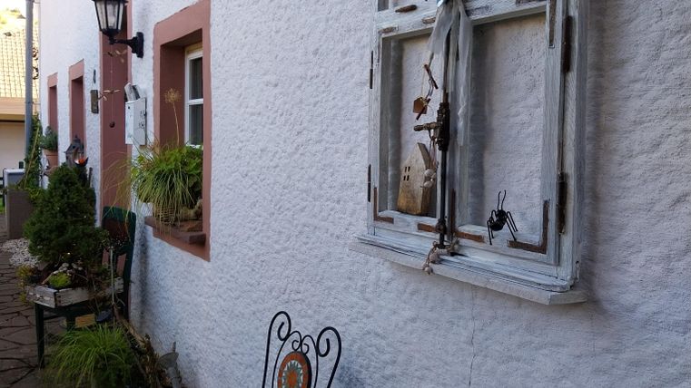 Een gezellige buitenruimte met een ronde tafel en twee stoelen. Aan de muur hangt een oud raam met gedroogde planten.