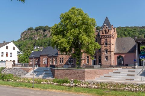 Außenfassade des Bahnhofs Gerolstein hinter einem dicht bewachsenen Baum, vor den Dolomitfelsen und am Ufer des Flusses Kyll.