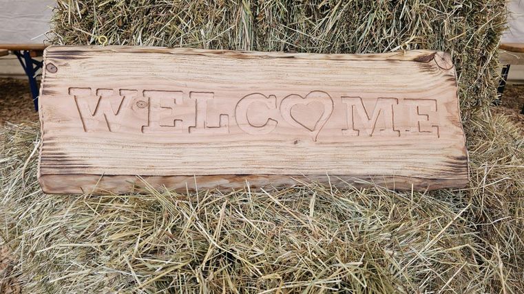 Ein Holzschild mit der Aufschrift "WELCOME". Im Hintergrund ist Heu sichtbar.