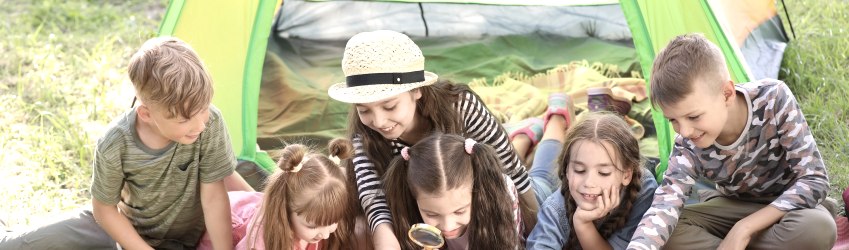 Zes kinderen liggen en zitten dicht tegen elkaar aan voor een kleurrijke tent op een weiland en bekijken een kaart die voor hen ligt. Sommige kinderen wijzen naar de kaart en één kind bekijkt de kaart nauwkeuriger met een vergrootglas.
