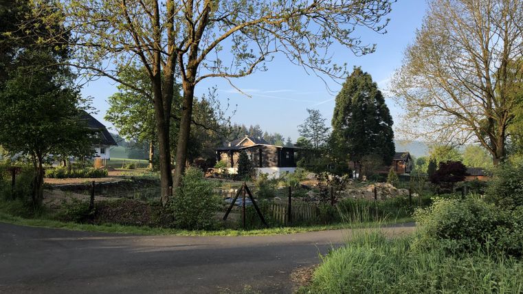 Een landelijke scène met bomen en een zachte weg. Op de achtergrond zijn gebouwen en een omheind gebied te zien.