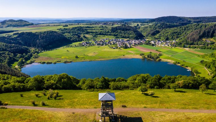 Eine schöne Aussicht auf einen See umgeben von grünen Wiesen und Hügeln. Im Hintergrund ist ein kleines Dorf zu sehen.
