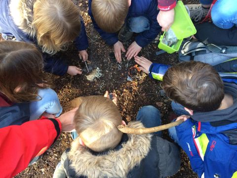 Eine Kindergruppe steht eng in einem Kreis zusammen. Akke Kinder blicken auf den Boden, wo einige der Kinder versuchen ein Feuer zu erzeugen.