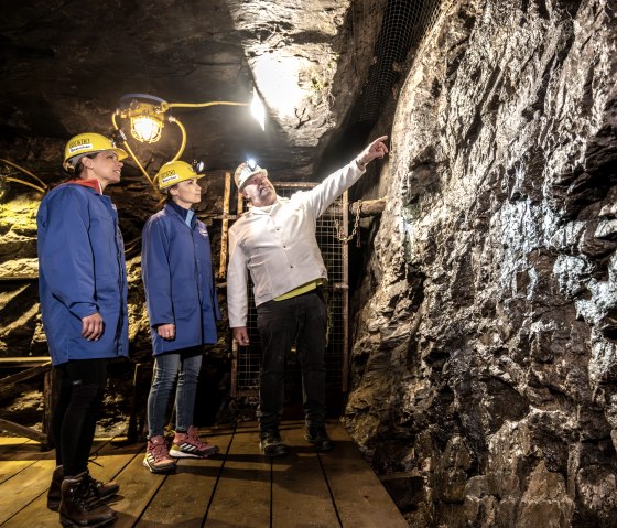Drei Personen in Schutzkleidung stehen in einem Bergwerk. Eine Person zeigt auf die Wand, die von einer Lampe beleuchtet wird., © Eifel Tourismus GmbH, Dominik Ketz
