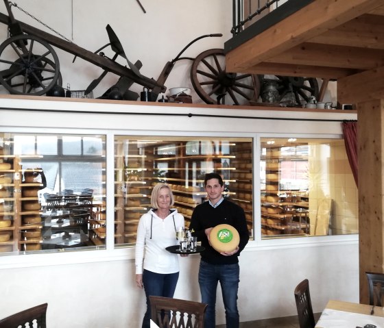 A man and a woman stand in front of a large glass pane, behind which numerous cheese wheels are maturing on shelves. The woman is carrying a tray with drinks and the man is carrying a cheese wheel.