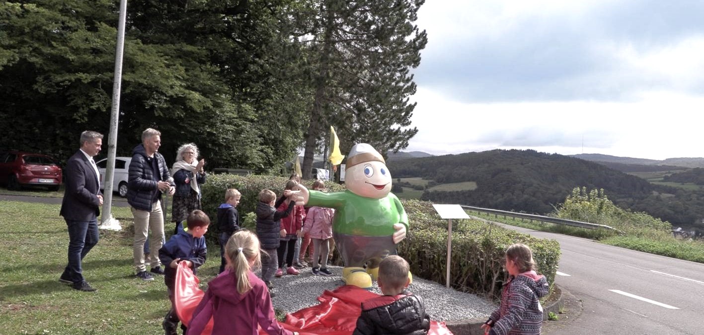 Standbeeld van mascotte Willi Basalt met groen shirt, grijze broek en gele schoenen wijst naar links. Enkele kinderen in een cirkel rond de figuur.