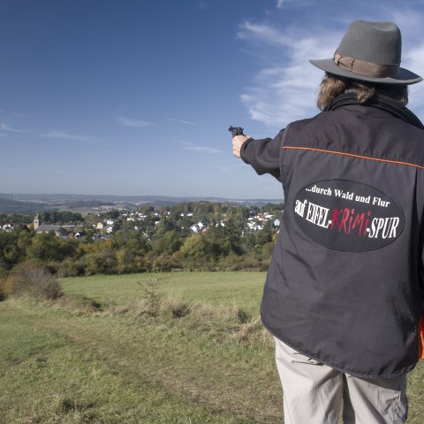 Person mit Hut und Jacke mit Aufschrift 'auf EIFEL KRIMI SPUR' zeigt auf eine weite Landschaft mit einem Dorf im Hintergrund., © Kappest