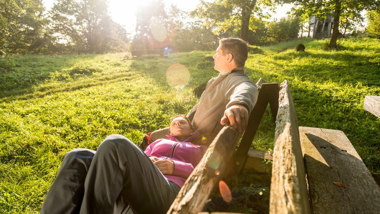 Un homme et une femme sur un banc dans une pelouse verte et luxuriante entourée d'arbres. La femme est allongée sur le dos, la tête sur les genoux de l'homme.