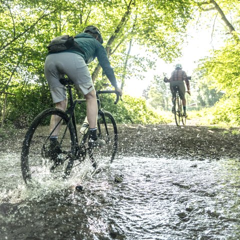 Ein Radfahrer auf einem Gravelbike fährt mitten durch eine große Fütze in einem Wald hindurch. Das Wasser spritzt zu allen Seiten. Vor ihm fährt ein weiterer Radfahtrer aus dem Wald heraus.