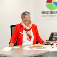 Eine Frau sitzt an eine Schreibtisch in einem hellen Büroraum mit Computer, Telefon und Tastatur vor sich und lächelt in die Kamera. Hinter ihr sind Prospektregale der Tourist-Information und das Logo der Touristik GmbH Gerolsteiner Land zu sehen.