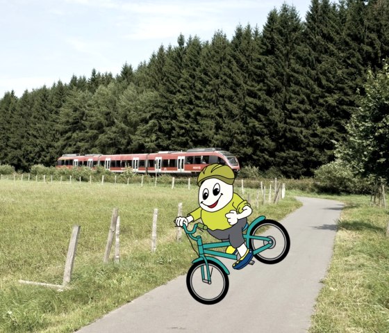 Stripfiguur Willi Basalt met een groen shirt, grijze broek, gele schoenen en een fietshelm op zijn hoofd zit op een fiets en rijdt over een fietspad. Op de achtergrond rijdt een trein.