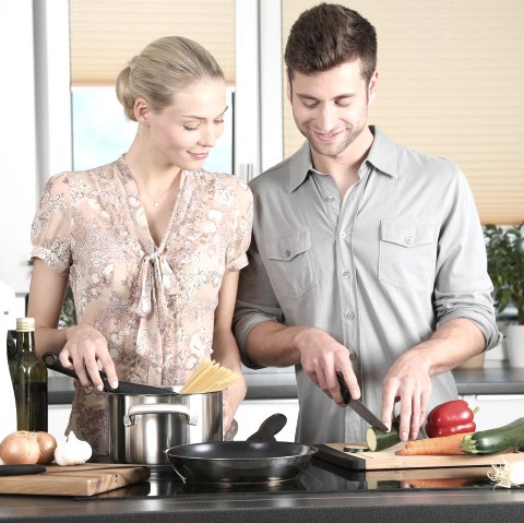 A couple stands side by side in the kitchen preparing a meal. Spaghetti is visible in the pot in front of them and vegetables are being chopped. 