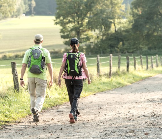 Zwei Personen wandern auf einem ländlichen Weg in der Eifel. Sie tragen Rucksäcke und sind von grüner Landschaft umgeben., © Eifel Tourismus GmbH, Dominik Ketz
