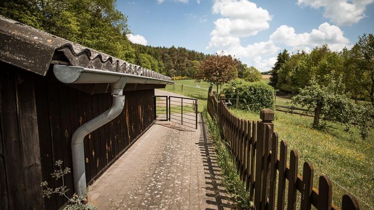 Ein Weg zu einem Holzgebäude mit einem Regenrinne. Im Hintergrund sind grüne Wiesen und Bäume unter einem blauen Himmel mit Wolken.
