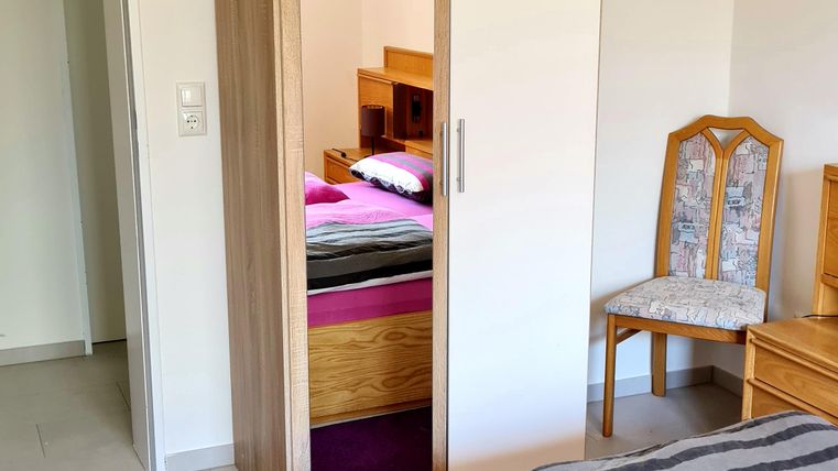 A simple bedroom with a bed, a wardrobe, and a wooden chair. The walls are light and the floor is tiled.
