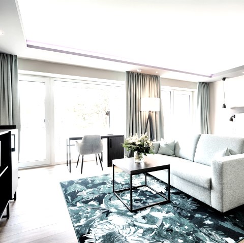 Modern eingerichtetes Hotelzimmer mit einem gro&szlig;en Doppelbett,  davor eine Couch auf einem Teppich mit Tisch und einem gro&szlig;en Flachbildfernseher an der Wand. Vor der gro&szlig;en Fensterfront steht ein Tisch mit einem Stuhl.