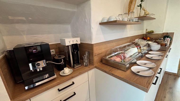 Eine moderne Küche mit einer Kaffeemaschine und einer Teestation. Auf der Ablage stehen Teller und frische Lebensmittel in einer Glasvitrine.