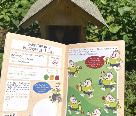 Wanderpass für den Barfußpfad im Bolsdorfer Tälchen mit Illustrationen und Infos zur Wanderung, gehalten vor einem kleinen Holzhaus im Grünen., © Touristik GmbH Gerolsteiner Land, Leonie Post
