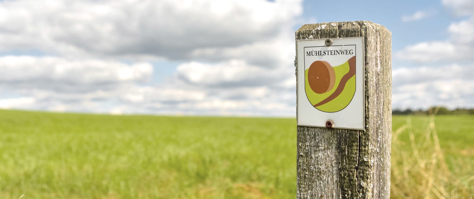 Signpost 'Mühlsteinweg' on wooden post in front of green meadow and cloudy sky., © Thomas Hendele