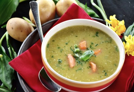 A bowl of potato soup on a saucer with a spoon, garnished with parsley, basil and carrot pieces. Next to it are potatoes and flowers.