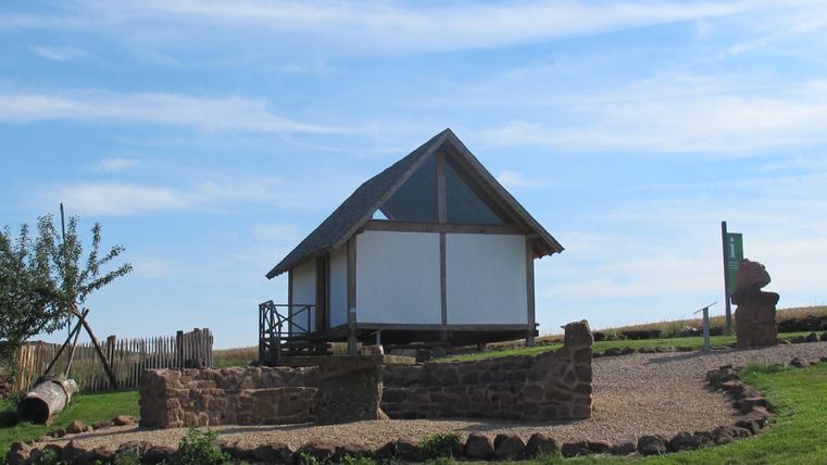 Ein kleines Fachwerkhaus steht auf einem Hügel. Im Vordergrund sind ein Baum, ein Zaun und ein Weg aus Steinen zu sehen. Der Himmel ist blau mit wenigen Wolken.