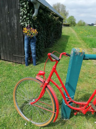 A red bicycle stands beside a green post on a meadow. In the background, a barn and a figure with flowering plants can be seen.