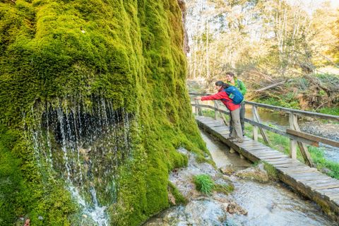 Zwei Wanderer stehen am Wasserfall Dreimühlen und strecken die Hand aus um das Wasser zu berühren.