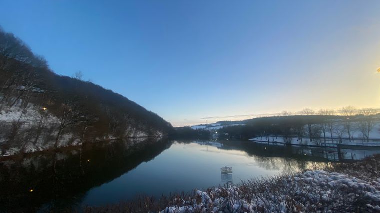 Ein See umgeben von verschneiten Bäumen und Sträuchern. Die Sonne verschwindet hinter dem Horizont.