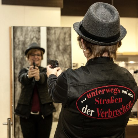 A woman stands in front of a mirror and points a gun at her own reflection. She is wearing a grey hat and a jacket with the inscription "...on the streets of crime".