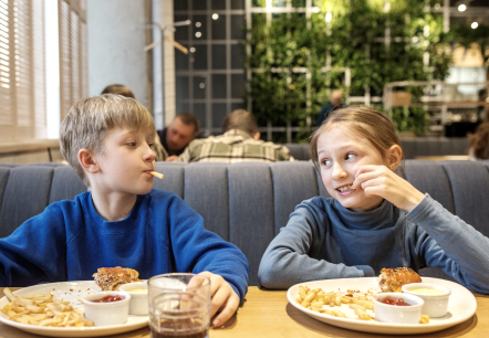 Two children sit next to each other at a table in a restaurant, eating French fries and a hamburger.