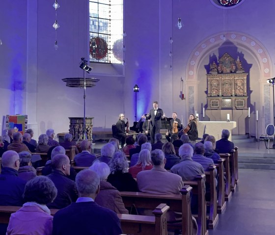 Die Besucher des Konzerts schauen alle nach vorne, wo der Dirigent und Pianist eine Ansprache h&auml;lt.