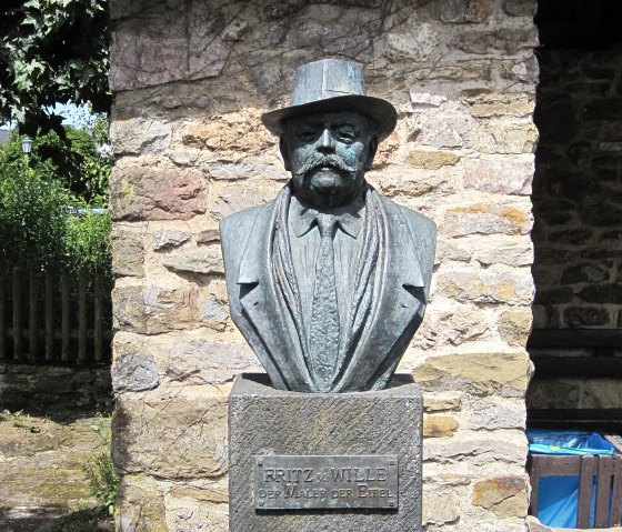 Bust of Fritz von Wille in front of a stone wall in Kerpen. The statue shows a man with a hat and moustache., © Touristik GmbH Gerolsteiner Land, Ute Klinkhammer