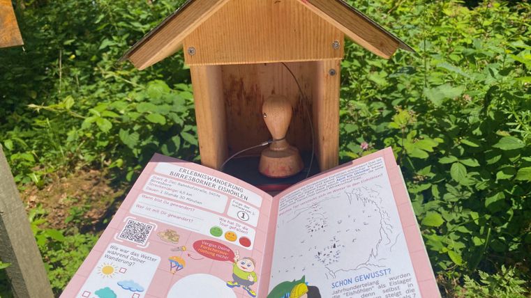 Ein geöffneter Wanderpass liegt vor einem Holzstempelhäuschen im Grünen.