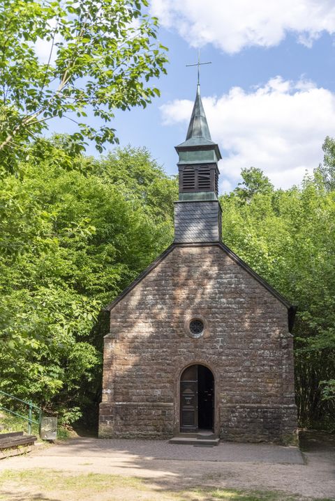 Steinkapelle mit spitzem Turm, umgeben von grünen Bäumen und blauem Himmel.