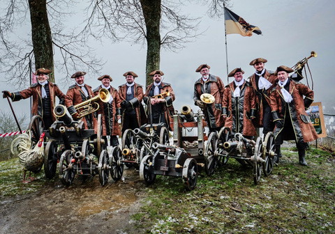 Eine Gruppe von Männern posiert  in historischer Kleidung, umgeben von kleinen Kanonen und Musikinstrumenten, vor dem Hintergrund einer trüben, winterlichen Landschaft.
