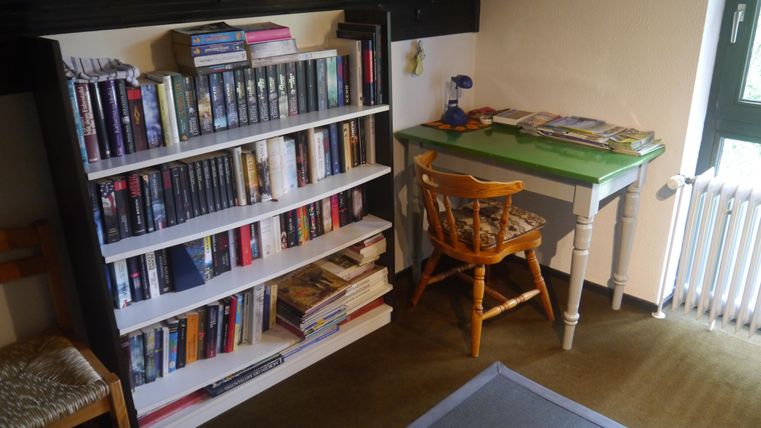 Ein gemütliches Zimmer mit einem Bücherregal und einem kleinen Schreibtisch. Der Schreibtisch ist grün und mit verschiedenen Büchern und einem Lampen ausgestattet.