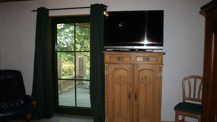 Een gezellige woonkamer met een televisie op een houten kast.
Ramen met uitzicht op groen en een blauwe bank in de hoek.