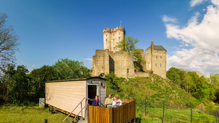 Eine alte Burg steht majestätisch auf einem Hügel, umgeben von Bäumen. Im Vordergrund sitzt eine Gruppe von Personen auf einer Terrasse eines Wohnwagens.