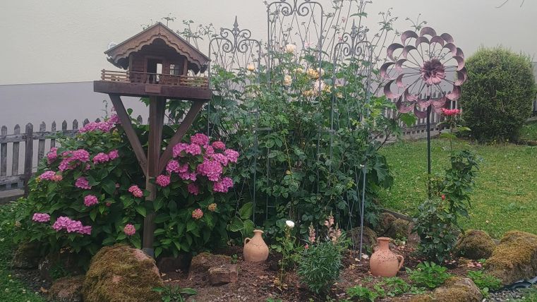 Ein kleiner Garten mit bunten Blumen, darunter rosa Hortensien und einer Vogelhaus-Dekoration. Im Hintergrund sind auch einige Kürbis-Pflanzen und Tontöpfe sichtbar.
