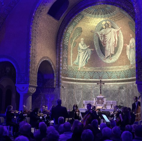 Schwach beleuchteter Innenraum der Erlöserkirche Gerolstein mit goldener Mosiakverzierung. Im Altarraum Musikanten und ein großes Publikum in den Sitzbänken.