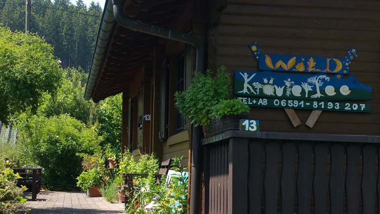 Ein gemütliches Holzhaus mit einem begrünten Vorgarten. Das Schild zeigt den Namen "Wild KräuterFee" und die Telefonnummer.