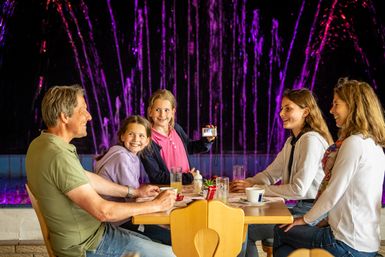 Eine Familie sitzt an einem Tisch und schaut auf die beleuchtete Wasserspielorgel.