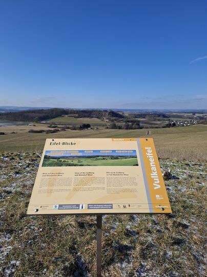 Eine Aussichtstafel steht auf einem Hügel mit Blick auf eine weitläufige Landschaft. Der Himmel ist klar und blau, und die Umgebung ist grün und sanft hügelig.
