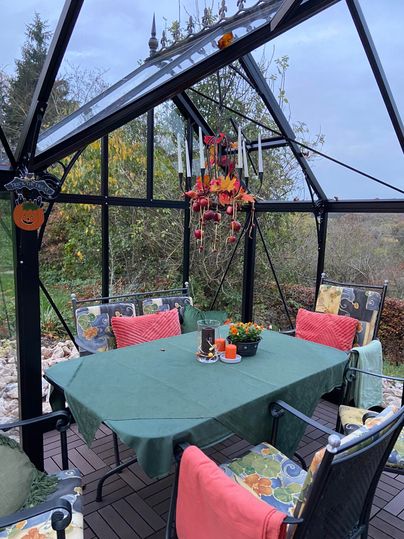 Ein gemütlicher Gartenpavillon mit einem grünen Tisch und bunten Stühlen. Auf dem Tisch stehen Blumen und Kerzen, umgeben von einer schönen Gartenlandschaft.