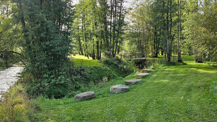 Grüner Park mit Bäumen, einem kleinen Fluss und einer Brücke im Kurpark Stadtkyll. Steine säumen den Weg entlang des Wassers.