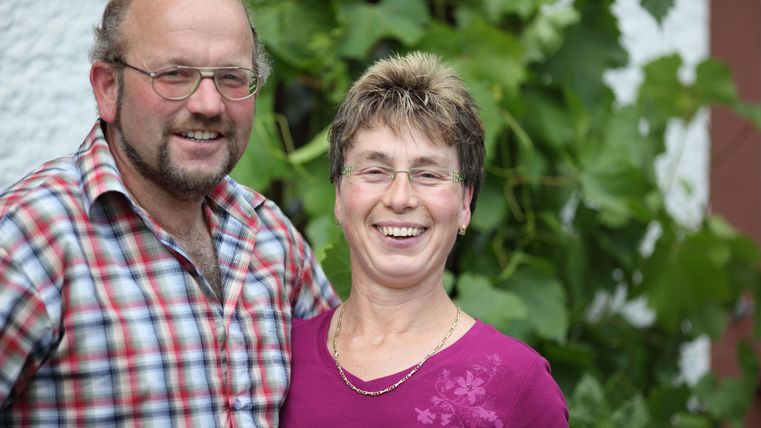 Ein freundliches Paar steht lächelnd vor einem grünen Hintergrund mit Weinblättern. Beide tragen legere Kleidung und strahlen Freude aus.